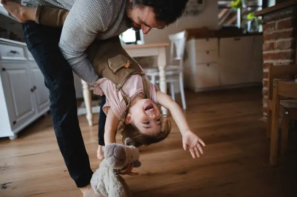 Photo of Mature father with small daughter playing indoors at home, holding her upside down.