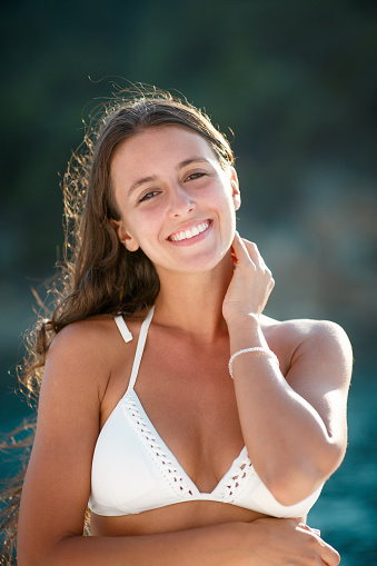 Portrait of beautiful young woman with long brown hair wearing white bikini and smiling at camera, touching neck with hand