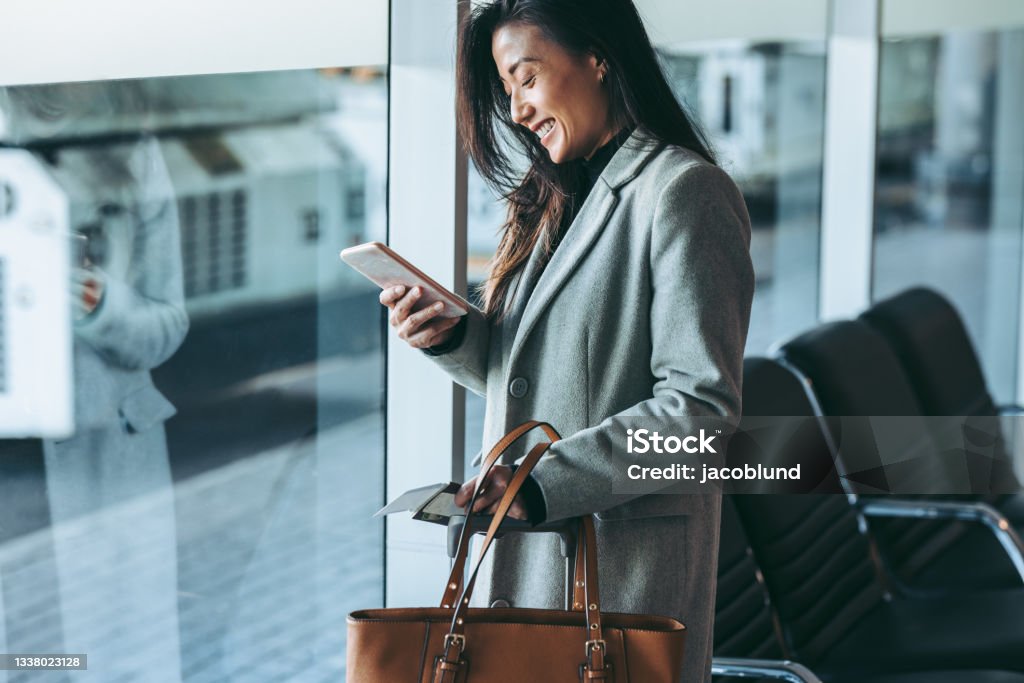 Business traveler waiting for her plane at airport Woman standing at airport lounge looking at her mobile phone and smiling. Business traveler at airport waiting for the flight. Business Travel Stock Photo