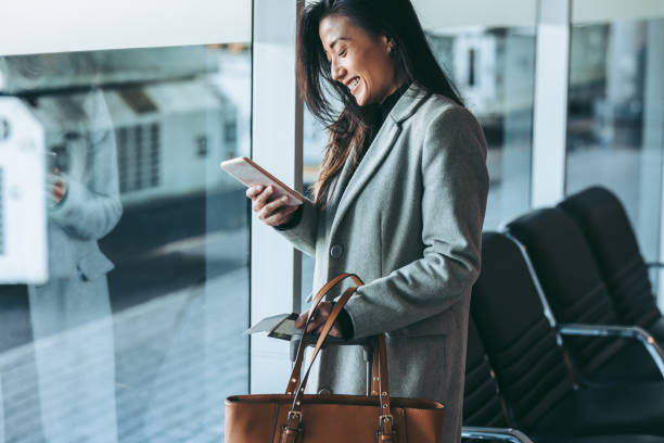 Business traveler waiting for her plane at airport Woman standing at airport lounge looking at her mobile phone and smiling. Business traveler at airport waiting for the flight. business travel stock pictures, royalty-free photos & images