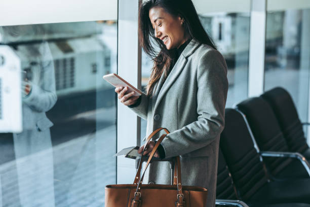 Business traveler waiting for her plane at airport Woman standing at airport lounge looking at her mobile phone and smiling. Business traveler at airport waiting for the flight. woman travel stock pictures, royalty-free photos & images