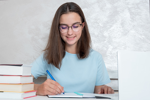 A smiling teenager student sitting at a table with books is writing an assignment in a notebook. The girl is a student studying for admission to the university, high school. Home schooling concept.