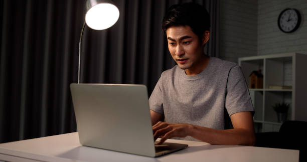 istock Attractive freelance programmer young Asian man working at home with laptop sitting alone at a desk in a dark room, Working late concept 1338027311