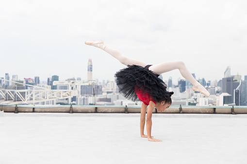 Asian ballerina ballet girl in red, black dress dancing on rooftop with cityscape view background. Young ballerina ballet girl dancing by stretching arms and leg on rooftop