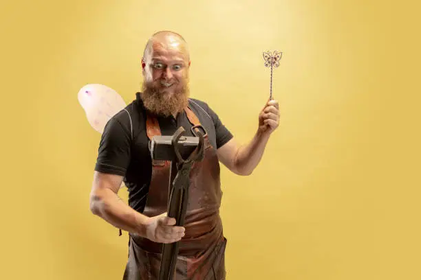 Photo of Comic portrait of muscular bearded bald man, blacksmith in leather apron or uniform isolated on yellow studio background. Concept of labor, retro professions, power, strength, humor