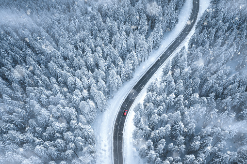 Red car driving on black asphalt road leading through snowcapped winter forest. Aerial view.