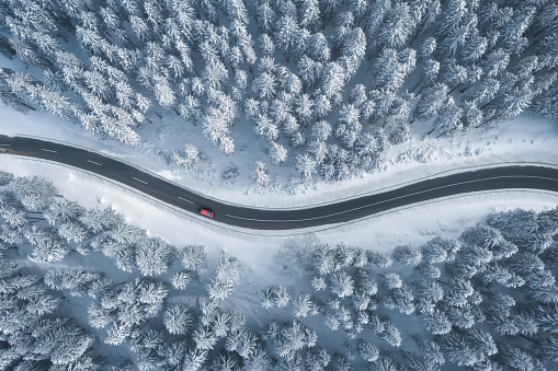 Red car driving on black asphalt road leading through snowcapped winter forest. Aerial view.