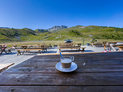 close-up of a cup of coffee with milk, on a wooden table in a terrace of a bar a summit of the Alps in the background the peaks of the mountains of the alps in summer, to the right the posts of the ski lift station sky. Cafe and table in the shade, the rest in the sun. the photo conveys tranquility, calm, spaciousness, health and beauty. Rest rest, quality of life