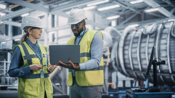 istock Two Diverse Professional Heavy Industry Engineers Wearing Safety Uniform and Hard Hats Working on Laptop Computer. African American Technician and Female Worker Talking on a Meeting in a Factory. 1338031146
