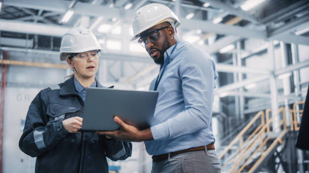 istock Two Diverse Professional Heavy Industry Engineers Wearing Safety Uniform and Hard Hats Working on Laptop Computer. African American Technician and Female Worker Talking on a Meeting in a Factory. 1338031147