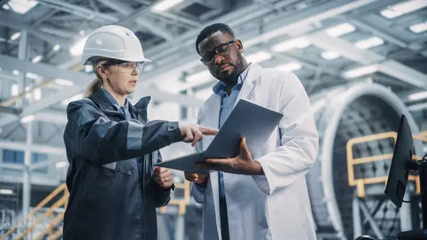 Photo of Team of Diverse Professional Heavy Industry Engineers Wearing Safety Uniform and Hard Hat Working on Laptop Computer. African American Technician and Female Worker Talking on a Meeting in a Factory.