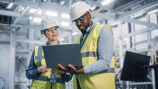 Team of Diverse Professional Heavy Industry Engineers Wearing Safety Uniform and Hard Hats Working on Laptop Computer. African American Technician and Female Worker Talking on a Meeting in a Factory.
