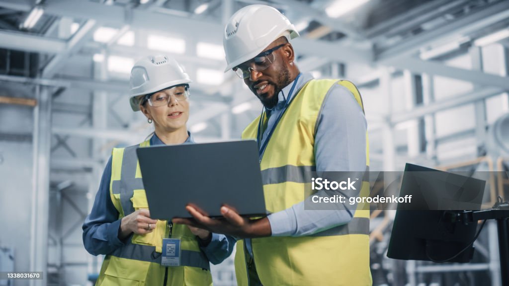Team of Diverse Professional Heavy Industry Engineers Wearing Safety Uniform and Hard Hats Working on Laptop Computer. African American Technician and Female Worker Talking on a Meeting in a Factory. Occupation Stock Photo