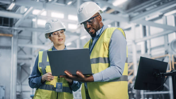 Team of Diverse Professional Heavy Industry Engineers Wearing Safety Uniform and Hard Hats Working on Laptop Computer. African American Technician and Female Worker Talking on a Meeting in a Factory. Team of Diverse Professional Heavy Industry Engineers Wearing Safety Uniform and Hard Hats Working on Laptop Computer. African American Technician and Female Worker Talking on a Meeting in a Factory. engineer stock pictures, royalty-free photos & images