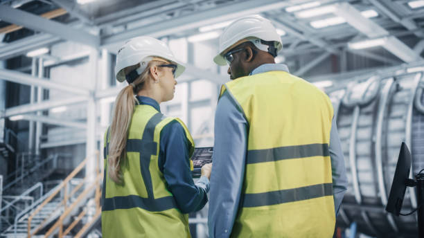 istock Two Diverse Professional Heavy Industry Engineers Wearing Safety Uniform and Hard Hats Working on Laptop Computer. African American Technician and Female Worker Talking on a Meeting in a Factory. 1338031673