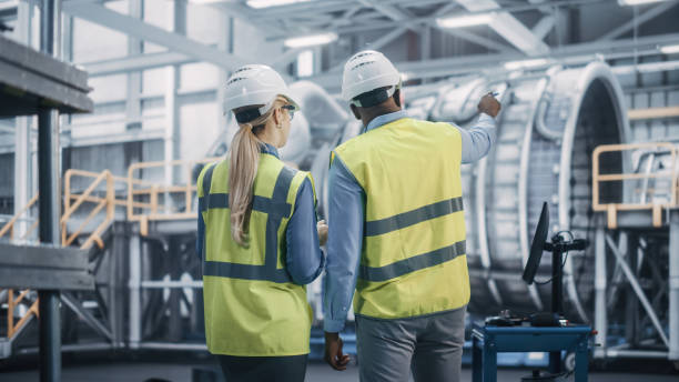 istock Two Diverse Professional Heavy Industry Engineers Wearing Safety Uniform and Hard Hats Working on Laptop Computer. African American Technician and Female Worker Talking on a Meeting in a Factory. 1338031682