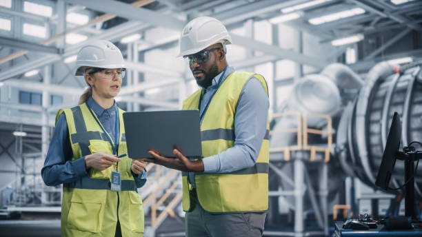 istock Two Diverse Professional Heavy Industry Engineers Wearing Safety Uniform and Hard Hats Working on Laptop Computer. African American Technician and Female Worker Talking on a Meeting in a Factory. 1338031699