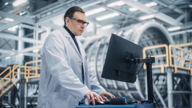 istock Professional Male Heavy Industry Engineer/Worker Working on a Computer. Confident Caucasian Industrial Specialist Wearing White Laboratory Coat and Safety Glasses in a Factory Facility. 1338031883