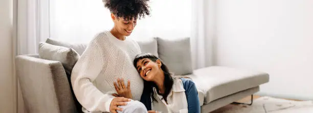 Lesbian mother-to-be listening to her unborn baby kicking. Happy young woman smiling cheerfully while listening to her wife's pregnant belly at home. Young lesbian couple expecting a baby.