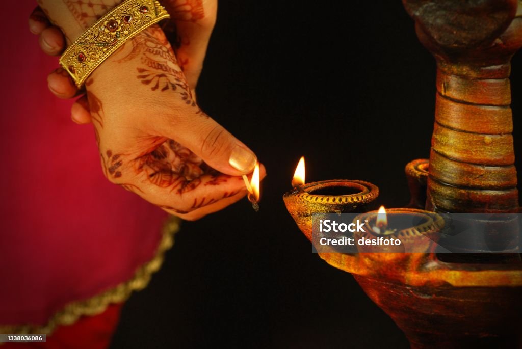Horizontal photograph of Indian Festival Diwali Deepawali being celebrated, a lady, woman with henna or mehndi and a big gold kangan lighting mitti or terracotta diya with matchstcick over black backgrounds Twof lit diyas , being lighted by a lady- concept of prayer and celebrations, symbol of peace and hope. Black background. A woman holding matchstick to one of mitti diya, earthen lamps with oil and jyot or lau. Apt for Diwali, Deepawali, Durga pooja, Ganesh Chaturthi Onam, Inauguration, opening of an event, Navratri festival related backdrops, wallpapers. The hand has beautiful mehendi pattern and a jadaau kangan. Navratri Stock Photo