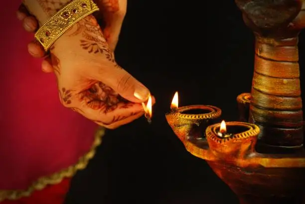 Twof lit diyas , being lighted by a lady- concept of prayer and celebrations, symbol of peace and hope. Black background. A woman holding matchstick to one of mitti diya, earthen lamps with oil and jyot or lau. Apt for Diwali, Deepawali, Durga pooja, Ganesh Chaturthi Onam, Inauguration, opening of an event, Navratri festival related backdrops, wallpapers. The hand has beautiful mehendi pattern and a jadaau kangan.
