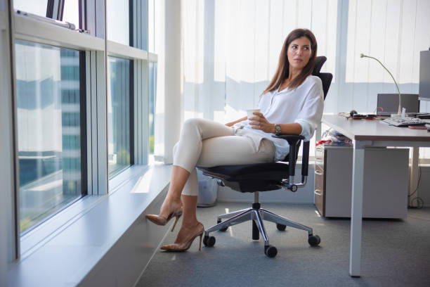 Businesswoman having a little break in her office chair, looking over shoulder with a frown Businesswoman sitting at her desk, holding paper coffee cup and looking over shoulder with a frown at someone interrupting her break woman sitting cross legged in chair stock pictures, royalty-free photos & images