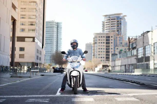 Photo of Wide shot of a young motorcyclist stopped at a traffic light in Barcelona. The man riding his scooter through the city on a large avenue lined with skyscrapers is waiting at the traffic light.