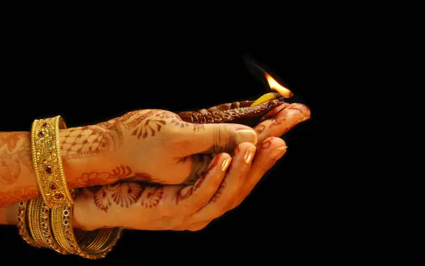 Photo of Diwali, Teej, Durga puja and Karwachauth celebrations themed festive picture of two  overlapping hands, with bangles in both hands and mehandi on palms holding a deepak or diya isolated over black backgrounds