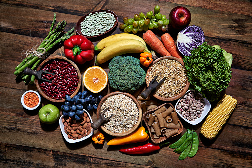 Dietary and healthy food themes. Table top view of fresh vegetables and legumes on rustic wooden table. Food is rich of fiber ideal for dieting and healthy eating. Includes corn, avocado, broccoli, orange fruit, grapes, bell pepper, lettuce, banana, apple  almonds and wholegrain pasta.