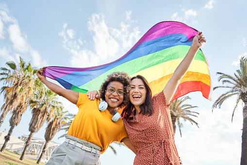 Young lesbian couple celebrates pride day looking at camera with rainbow flag. Concept of homosexuality, gay.