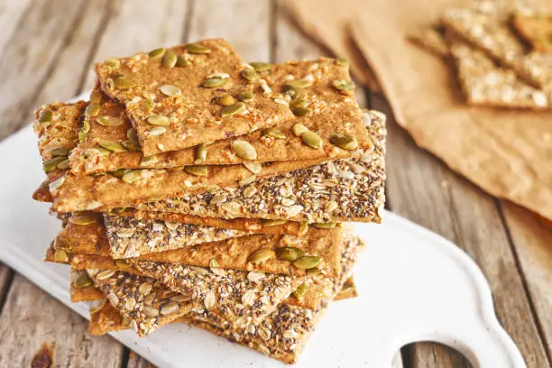 Crunchy crispbread on white ceramic cut board on wooden table. Whole Grain crisp bread with pumpkin, sunflower, chia, linen and sesame seeds. Healthy snack. Bio-organic dieting product.