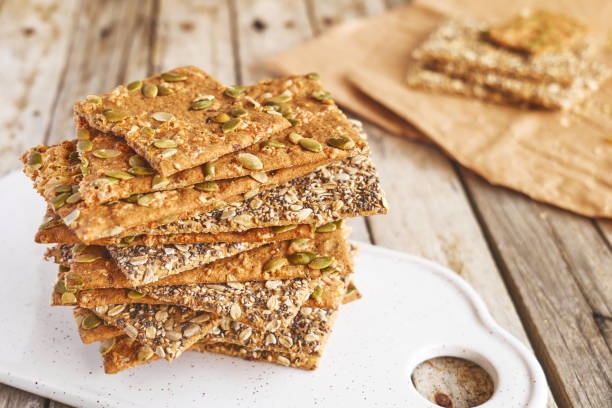 istock Crunchy crispbread on white ceramic cut board on wooden table. Whole Grain crisp bread with pumpkin, sunflower, chia, linen and sesame seeds. Healthy snack. Bio-organic dieting product. 1338068288