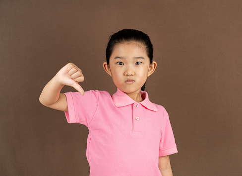 Baby girl giving thumbs down hand gesture, white background with copy space, close-up. Portrait unhappy child girl with long brown hair, wearing gray dress, showing dislike gesture