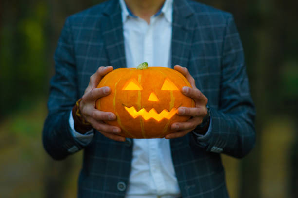 Man in suit holds a carved glowing pumpkin lantern with a creepy face. Halloween concept stock photo