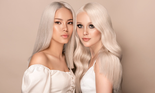 Double portrait of young attractive models with dyed blonde hairstyle. Group portrait of blondes with long, well cared hair. Sensual looks and elegant makeup. Coloration in blonde. Stylish blonde hair.Hairdressing art.