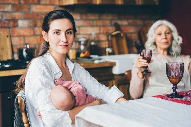 young woman breastfeeds baby and looking to the camera and sitting on wooden chair next to grey haired mother. stock photo