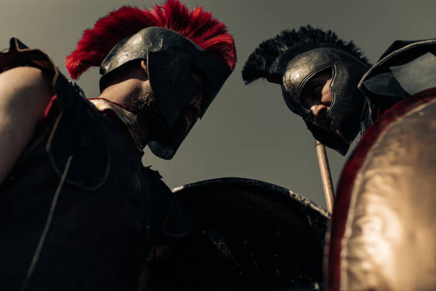 Duel between two ancient greek warriors in battle dress with shields and spears against sky background.