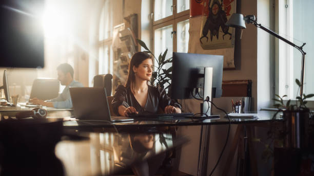 zwei stilvolle mitarbeiter arbeiten an computern in der kreativagentur im loft office. schöner manager, der korrespondenz-e-mails eingibt. sonniger renovierter raum mit pflanzen, künstlerischen postern und großen fenstern. - kleines büro stock-fotos und bilder