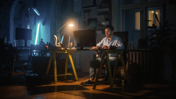 istock Focused and Productive Man Works on a Desktop Computer in Creative Agency in Dark Loft Office in the Evening. Renovated Stylish Decor with House Plants, Artistic Posters and Big Windows. 1338090457