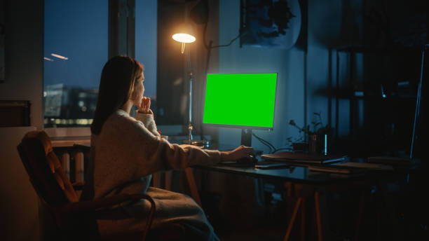 istock Female Manager Works on a Desktop Computer with Green Screen Chromakey Mock Up in Creative Agency in Dark Loft Office with Stylish Design in the Evening. Over the Shoulder Shot. 1338090470