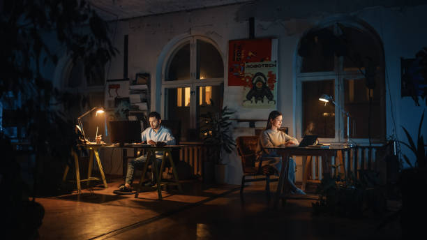 istock Two Stylish Employees Working on Computers in Creative Agency in Loft Office. Colleagues Answer Emails and Manage Marketing Projects. Dark Renovated Space in Evening with Plants and Artistic Posters. 1338090487