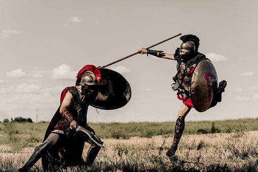 Battle with spear and sword between two ancient greek or roman warriors in battle dress and cloaks on meadow against sky background.