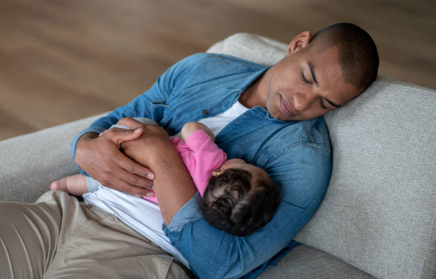 измученный отец спит на диване, держа на руках своего малыша - tired men african descent sadness стоковые фото и изображения