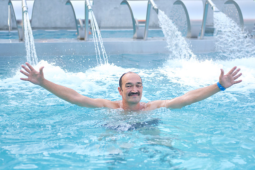 Smiling mature asian man looking at camera opening your hands in outdoor thermal pool with hydromassage over background of injectors with water jets. Physical health concept.