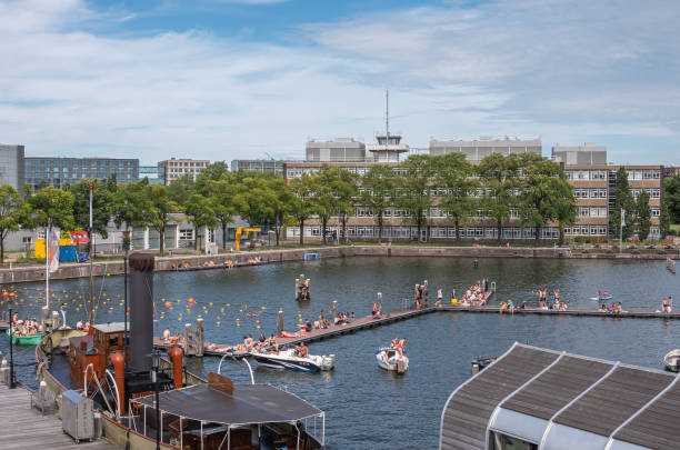 Amsterdam, Netherlands - August 15, 2021: Open water public swimming dock at Marineterrein adjacent to Maritime Museum under blue cloudscape with many people in and out of the water.