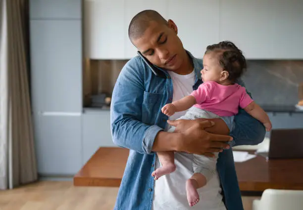 Photo of Busy father multi-tasking at home carrying the baby while talking on the phone