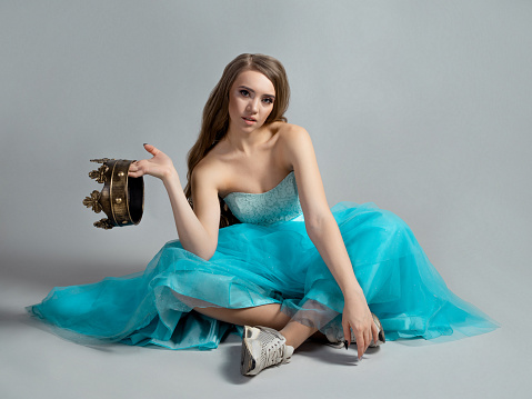 Modern princess, beauty in a crown and a lush blue dress and sneakers, bold contrast, sitting on the floor, gray background