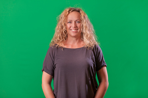 Front portrait of mature blonde woman on green background. She has grey T-shirt, happy smiling and looking at camera.