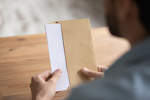 Man sit at table opens envelope with letter or post card inside, close up view over male shoulder. Paper correspondence with information, bank notification, paperwork at workplace, invitation concept