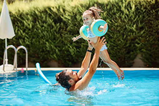 A father and his 4 year old girl playing together in a swimming pool. Dad throws the boy high up in the air and he lands with a big splash.